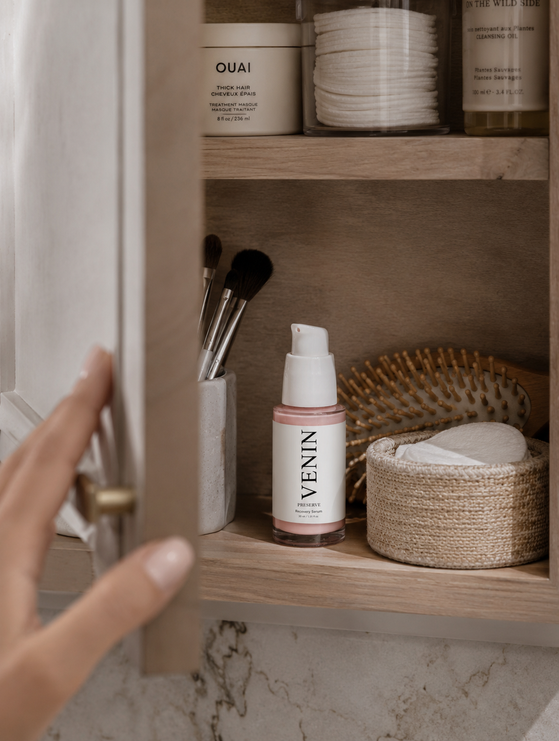 A hand opens a light wood bathroom cabinet door, revealing a Venin skincare pump bottle on a marble countertop alongside other bathroom items.