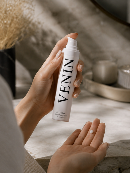 A person holds a white pump bottle labeled VENIN in front of their hand, with a small amount of cream visible on the palm, in a softly lit bathroom setting.