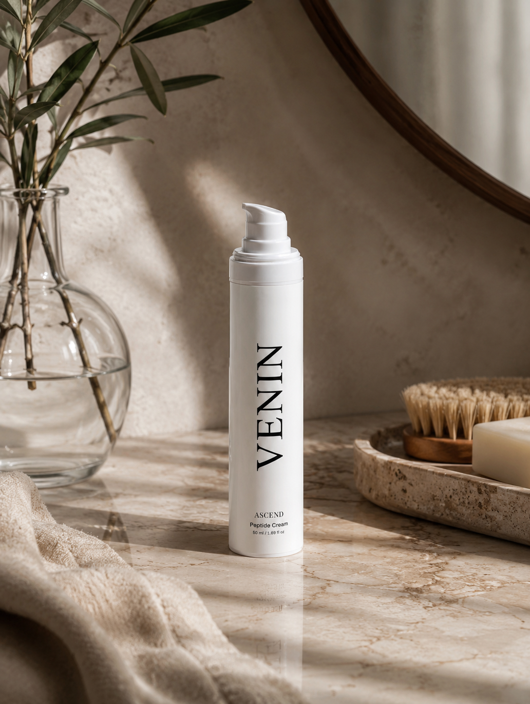 A white pump bottle of VENIN Peptide Cream stands on a marble countertop in a warm, sunlit bathroom setting with a glass vase, mirror, and soap brush nearby.