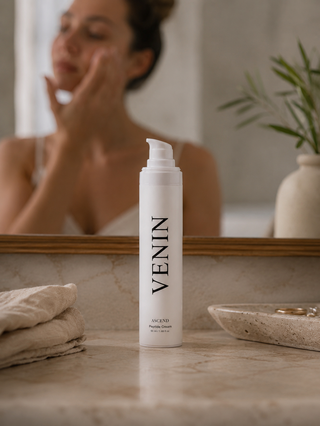 A white pump bottle of VENIN Peptide Cream stands on a marble countertop in a bathroom, with a blurred mirror reflection of a woman applying cream to her face in the background.