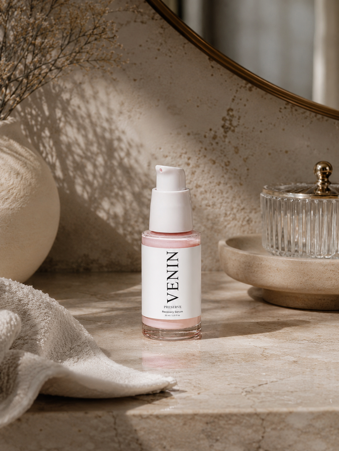 A bottle of VENIN Recovery Serum with a white pump and label, set on a beige stone surface next to a textured towel and decorative ceramic objects.