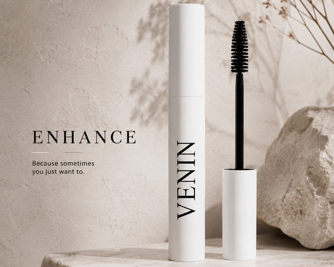 VENIN Enhance mascara tube in white with black lettering, shown next to the black mascara wand on a stone surface with a textured beige wall and dried floral decor.